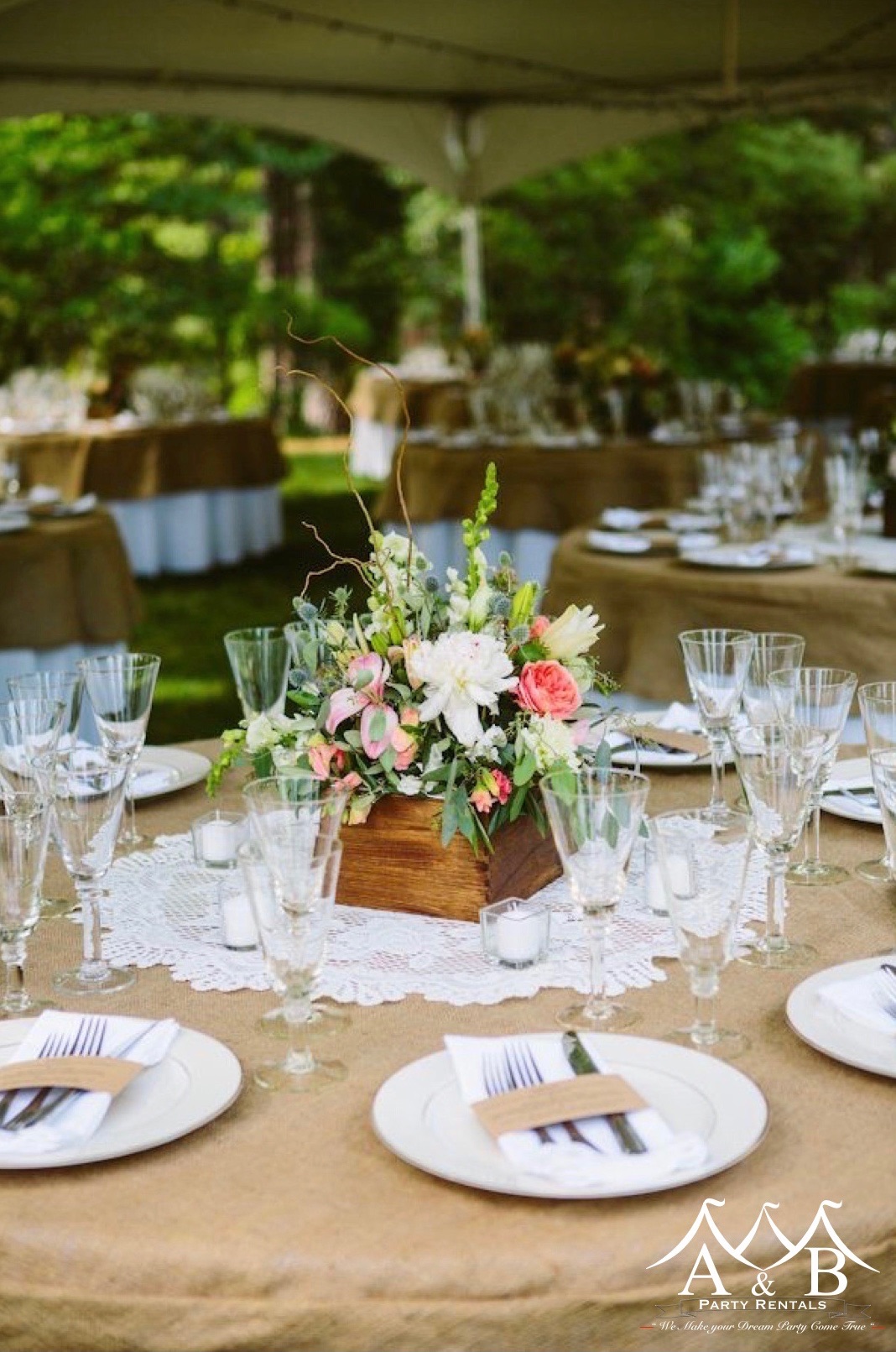 A close-up of a round table with wedding settings, a floral centerpiece, and various similar tables under a tent at a wedding. Image represents elegant table settings at a wedding event provided by A&B Party Rentals in Salisbury, MD.