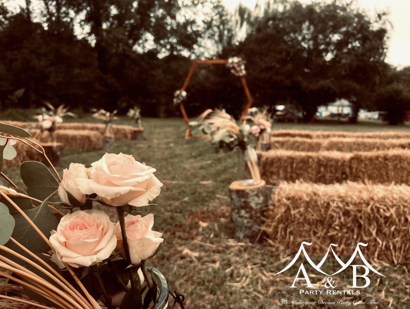 A cedar archway in the background of a wedding aisle outdoors, surrounded by rows of haystacks and flowers for a wedding. The image captures a scenic outdoor wedding setting with a cedar archway overlooking the wedding aisle, complemented by rows of haystacks and floral arrangements. This charming scene is perfect for outdoor weddings and is provided by A&B Party Rentals in Salisbury, MD.