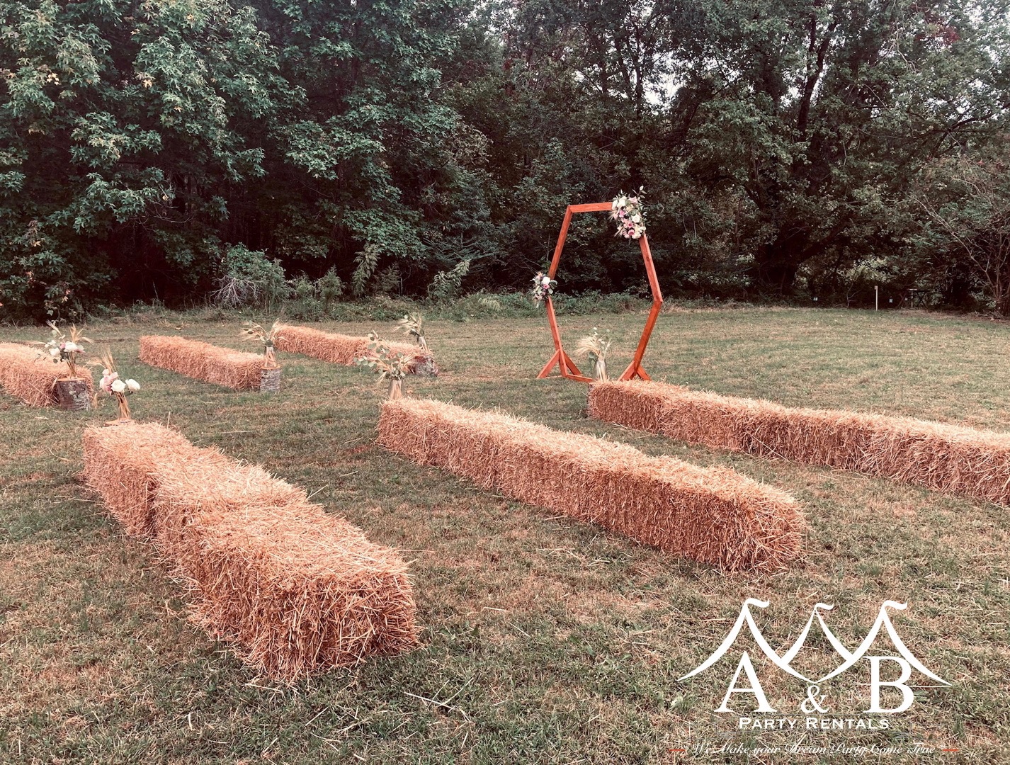A cedar archway designed for outdoor weddings set between rows of hay stacks. The image showcases a picturesque outdoor wedding setting with a cedar archway positioned between rows of hay stacks, providing a rustic and charming backdrop for ceremonies. Available through A&B Party Rentals in Salisbury, MD.