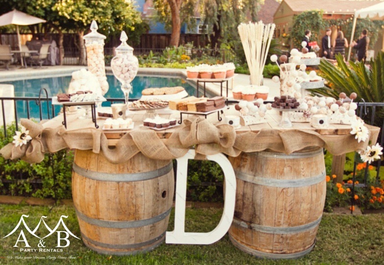 A wooden table placed on top of wooden barrels adorned with floral arrangements, wedding catering items, and linens, featuring a large marquee letter 'D' in the center. The image showcases a beautifully decorated table setting with a variety of wedding elements, including catering items and floral arrangements, complemented by a prominent marquee letter 'D'. Available through A&B Party Rentals in Salisbury, MD.