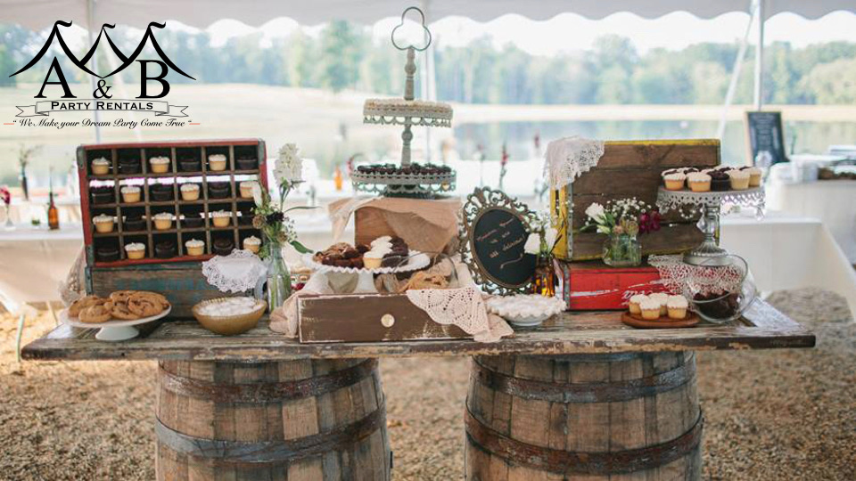 A wooden table placed on top of wooden barrels showcasing a rustic wedding snack spread. The image depicts a charming rustic setup with a variety of snacks on display, ideal for weddings and events. Available through A&B Party Rentals in Salisbury, MD.