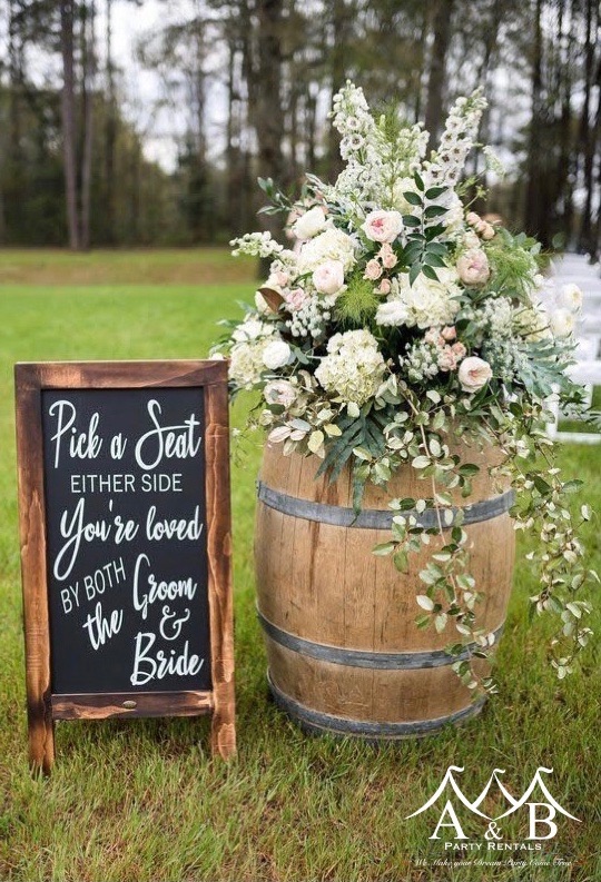 A floral arrangement outdoors on a long table with a chalkboard sign at a wedding. Image showcases outdoor wedding decor and floral arrangements provided by A&B Party Rentals in Salisbury, MD.