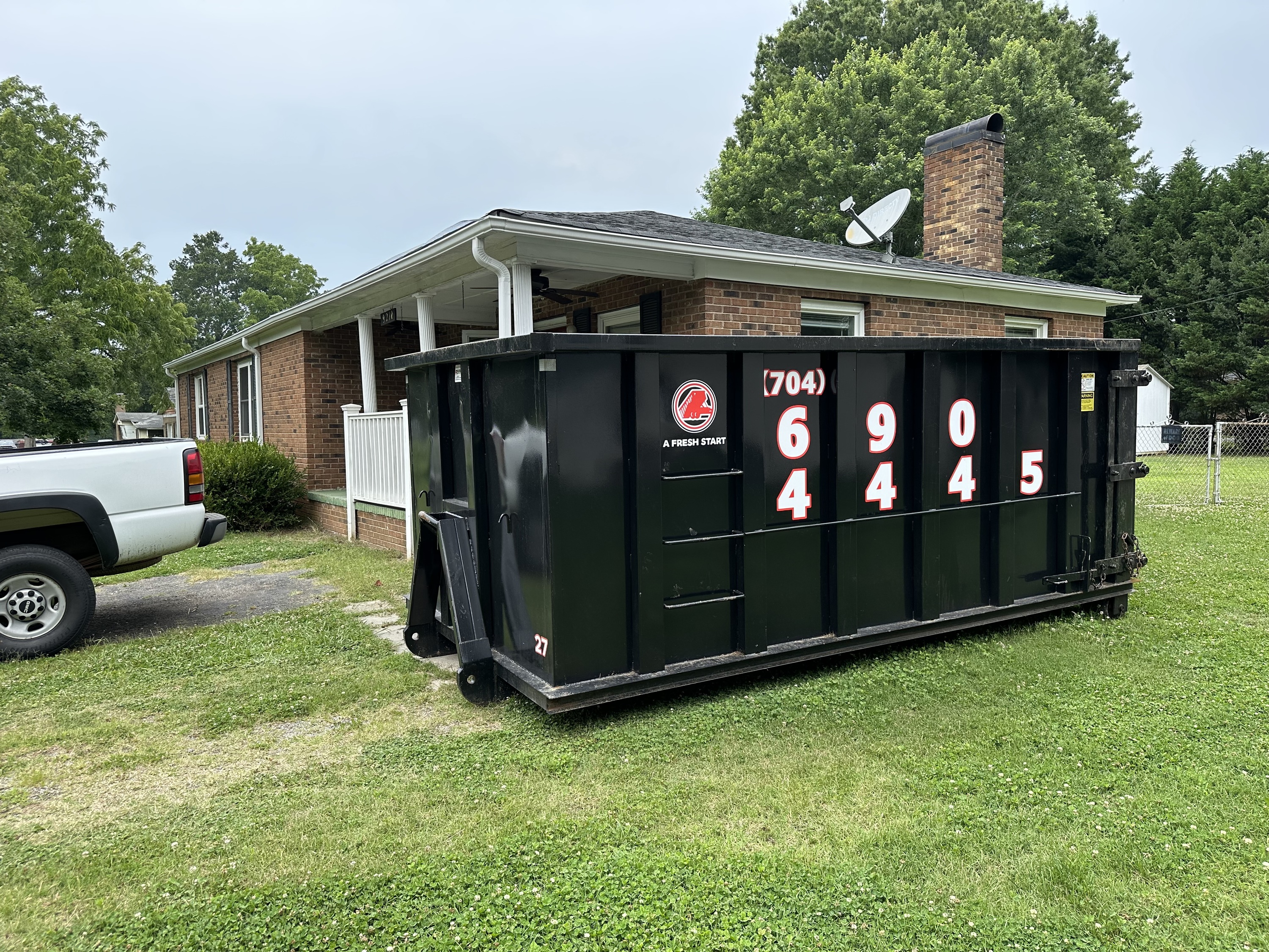 A black dumpster with a phone number printed on the side is placed on the grass beside a brick house and a white pickup truck.