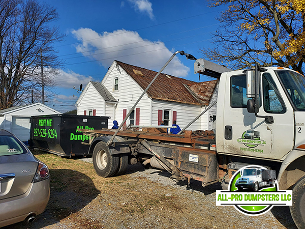 Image of a roll-off dumpster being used at a residential or commercial location, possibly with company personnel in the background.