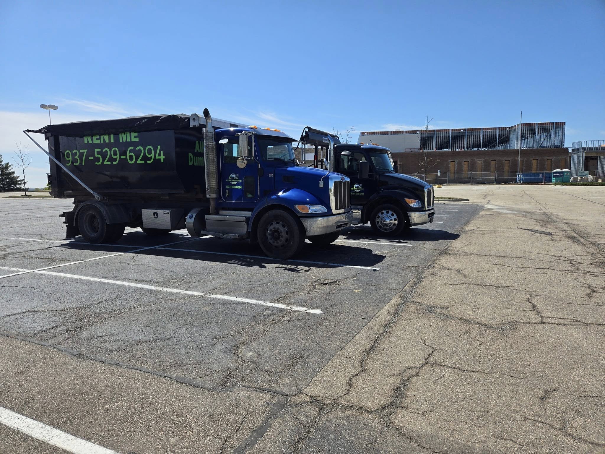 Two All-Pro Dumpsters roll-off dump trucks, one blue and one black, at a Industrial site.