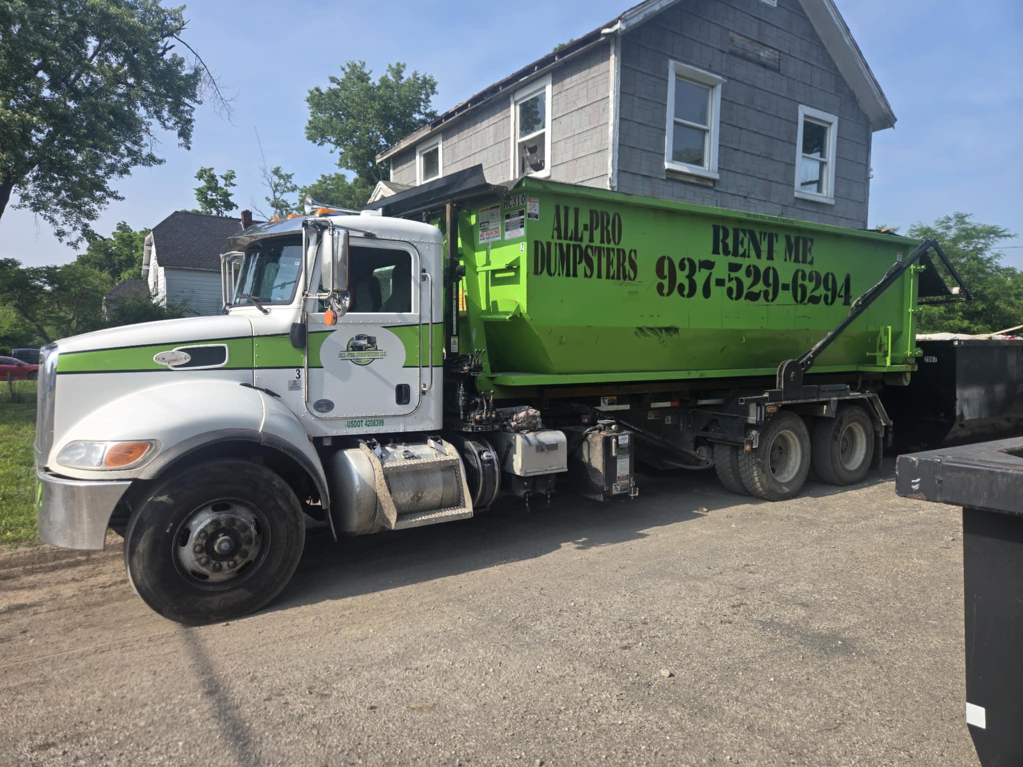 A white and green All-Pro Dumpsters truck delivering a large green dumpster to a house.