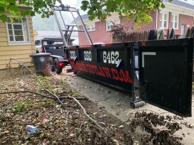 All Things Dumpster Rentals red dumpster being delivered on a trailer to a residential driveway beside a white house with a carport.
