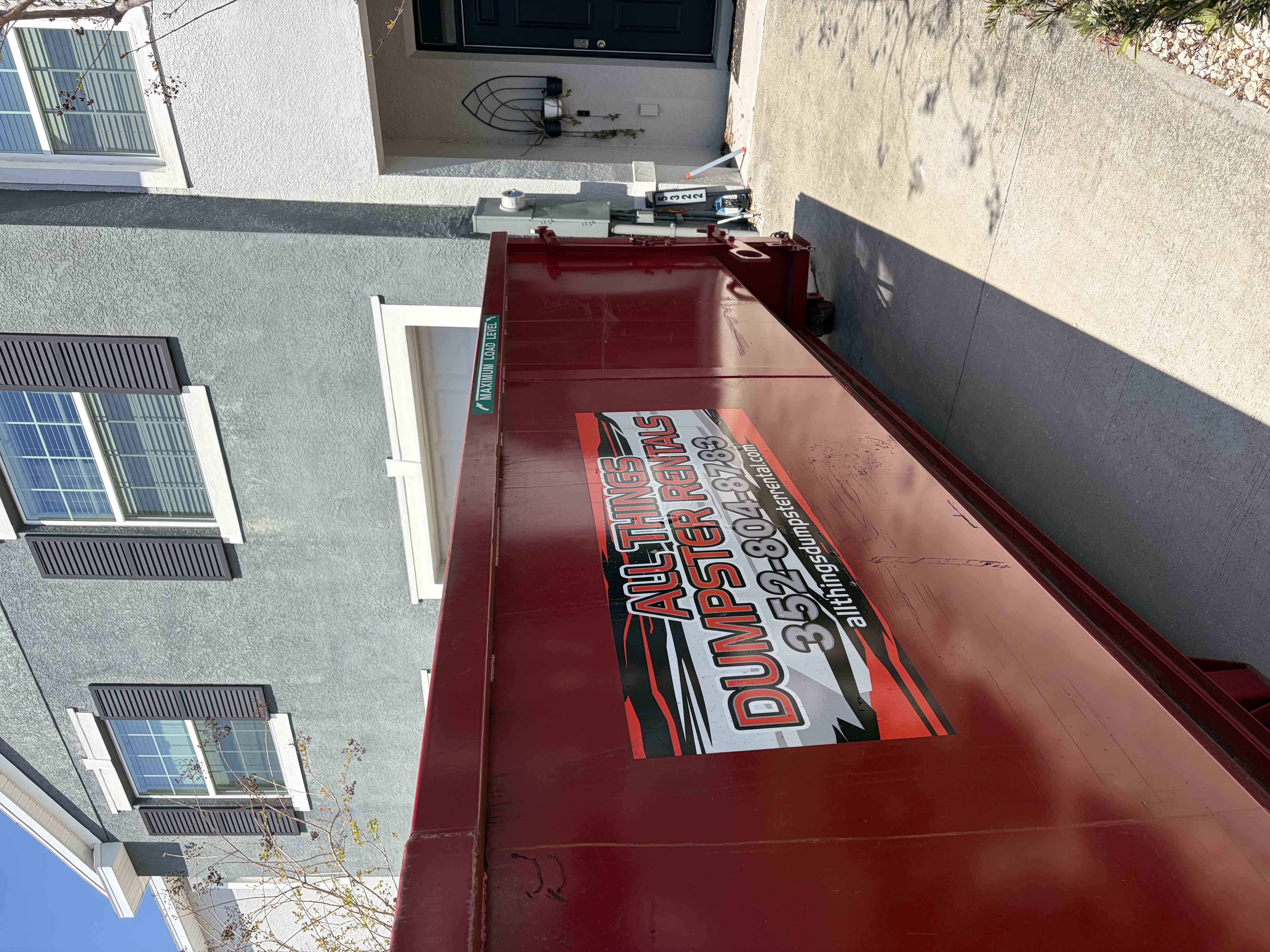 All Things Dumpster Rentals red dumpster positioned on a residential driveway in front of a house and garage.