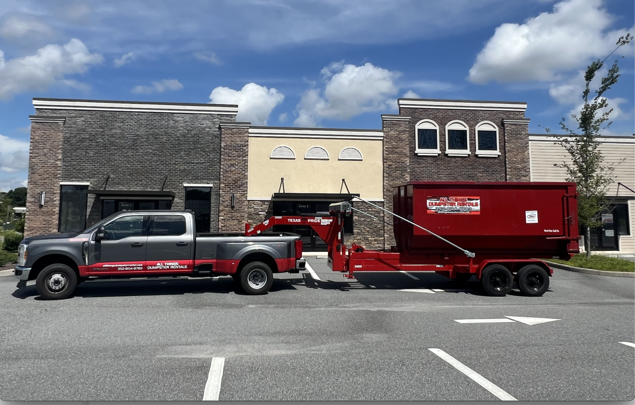 All Things Dumpster Rentals black pickup truck towing a red dumpster trailer in a parking lot.