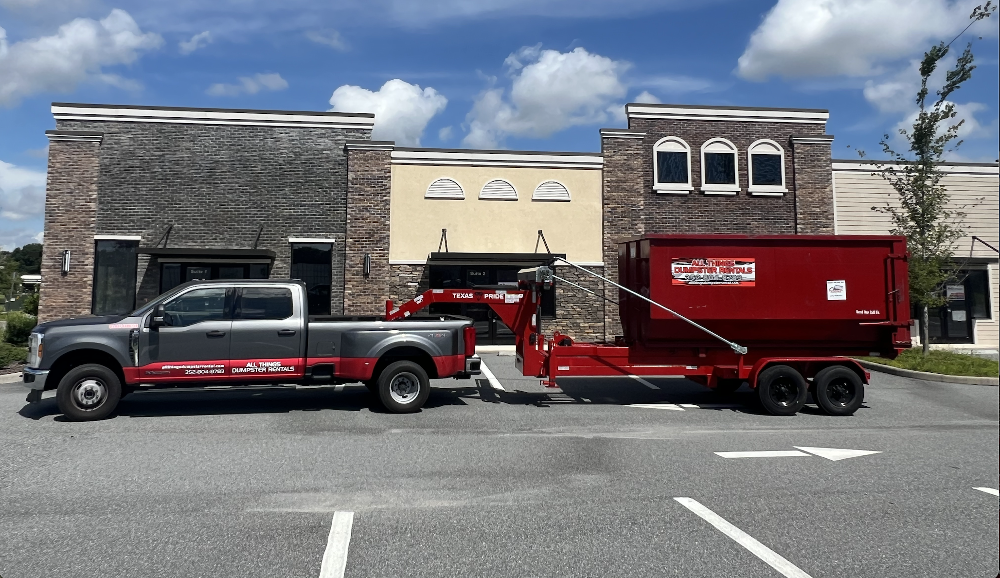 All Things Dumpster Rentals black pickup truck towing a red dumpster trailer in a parking lot.