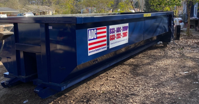 A roll-off dumpster from American Dumpster Company sitting on a dirt lot near trees