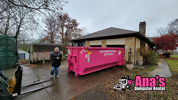 Large pink dumpster from Ana's Dumpster Rental placed on a residential driveway with a person standing nearby