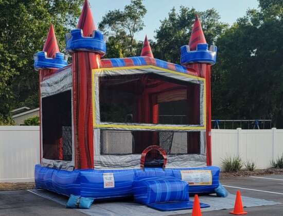 Red and blue inflatable bounce house castle with turrets set up in a parking lot, surrounded by orange safety cones