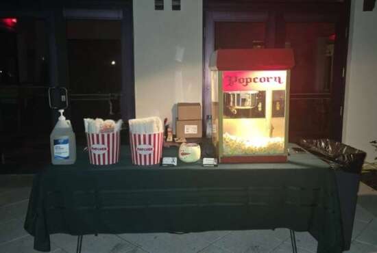 Illuminated popcorn machine with striped popcorn buckets and hand sanitizer set up on a black-draped table at a nighttime event