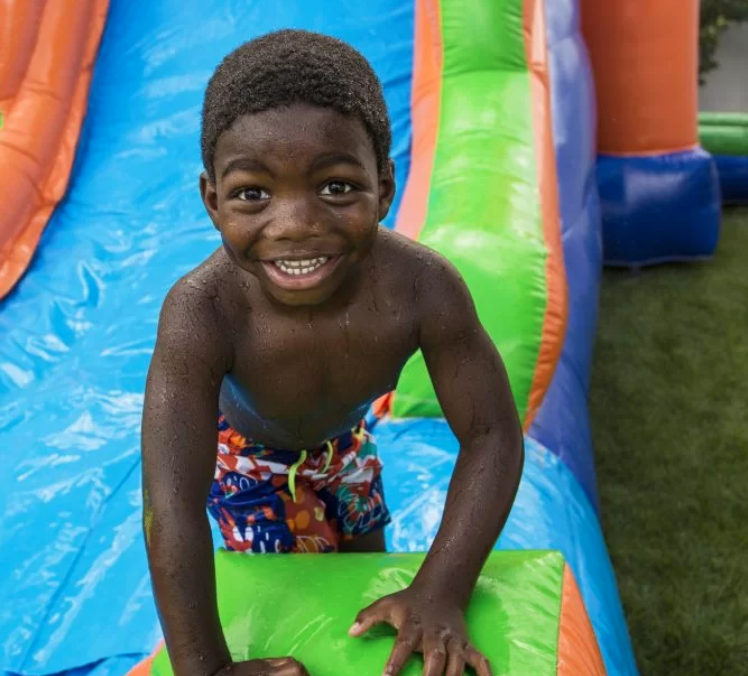 A happy child smiles while climbing on a colorful inflatable water slide with blue, green, and orange sections. This image highlights the clean, family-friendly water slide rentals A Party Solution provides for Cedar Hill, TX parties, church events, school fun days, and backyard celebrations.