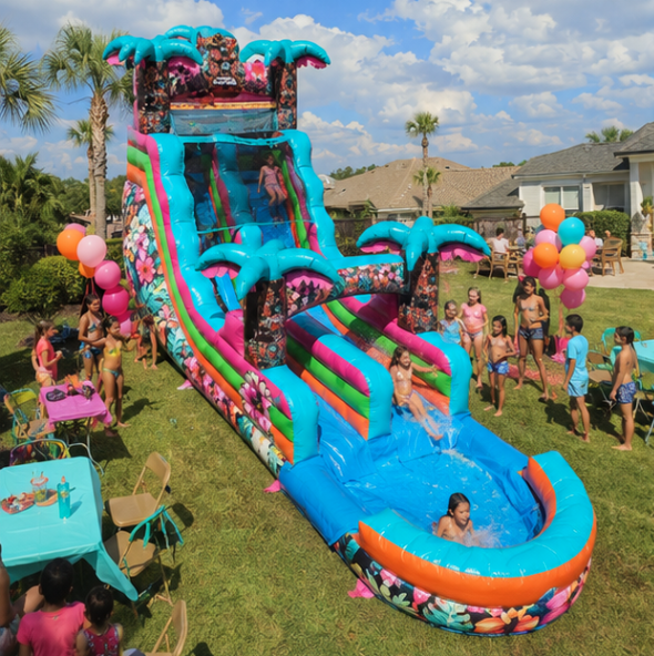 Pink, teal, and floral inflatable water slide with palm tree accents surrounded by children at a backyard celebration.