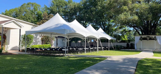 Outdoor wedding ceremony setup with white high peak tents and rows of black chairs on a green lawn