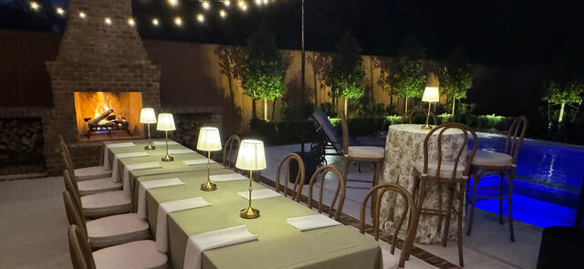 Outdoor dinner party with long tables, table lamps, fireplace, and pool at night