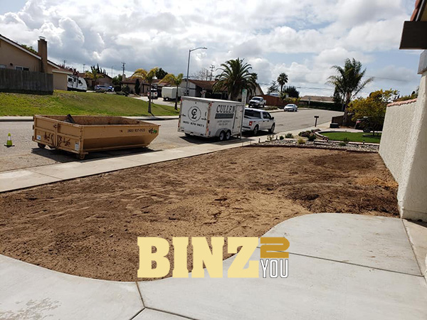 Residential yard with a BINZ 2 YOU dumpster and a trailer parked by the sidewalk. Residential yard with a BINZ 2 YOU dumpster and a trailer parked by the sidewalk.