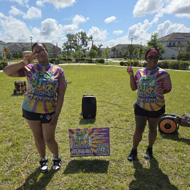 Two Bounce N Around Rentals event attendants in company shirts on a neighborhood green at an HOA event in Narcoossee, Saint Cloud, FL, beside a company yard sign, speaker and generator