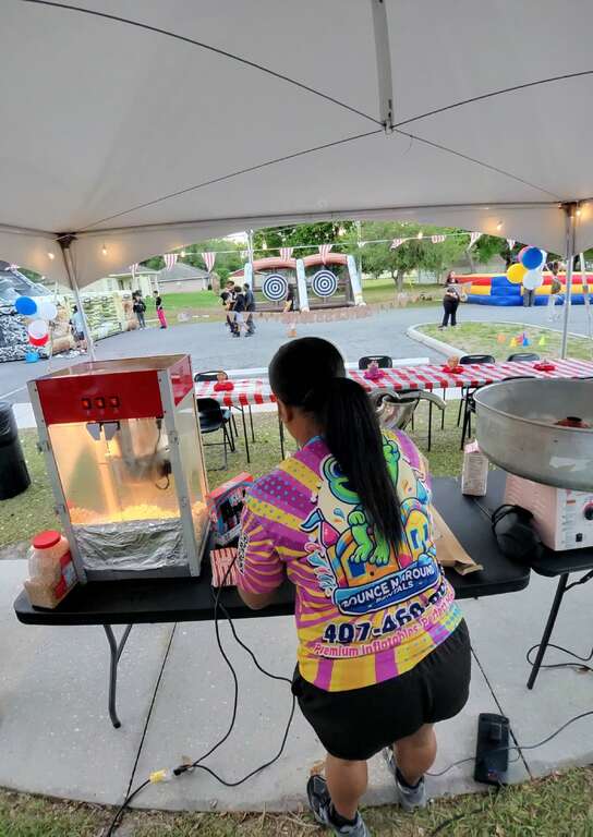 Bounce N Around Rentals staff member running a popcorn machine and a cotton candy machine under a high-peak tent at a church youth event in Saint Cloud, FL, with the Black Ops obstacle course, axe throw game and Joust arena behind