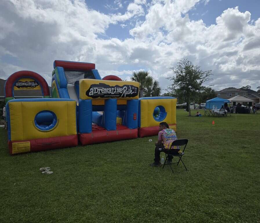 Adrenaline Rush 2 obstacle course on a grass field with a Bounce N Around Rentals staff member seated nearby at a Tivoli Reserve community event in Davenport, FL