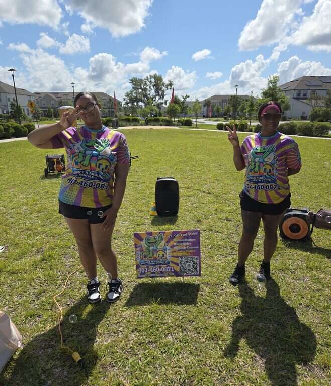 Two Bounce N Around Rentals staff members in company shirts beside a yard sign, Bluetooth speaker and generator at the Weslyn Park HOA event in Saint Cloud, FL