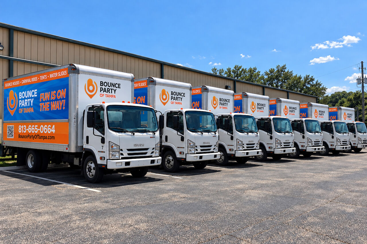 Seven white Bounce Party of Tampa box trucks in blue and orange wraps reading Fun is on the way!, parked in a row outside the warehouse
