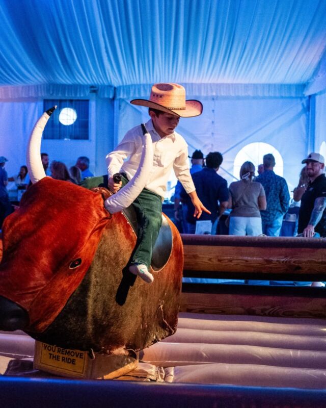 A boy in a cowboy hat riding a mechanical bull at an evening gala under a tent