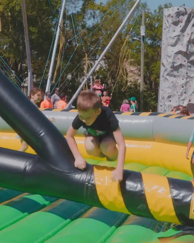A boy climbing over the arm of an inflatable sweeper game, with a bungee trampoline and a rock wall behind