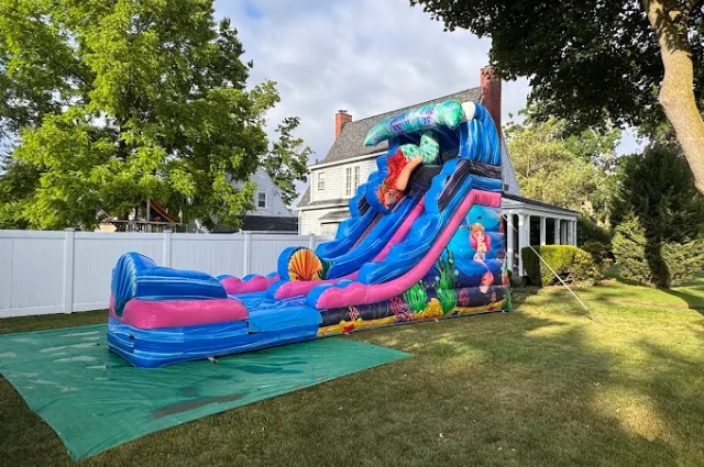 Large ocean-themed inflatable water slide with splash pool set up in residential backyard surrounded by trees and white fencing