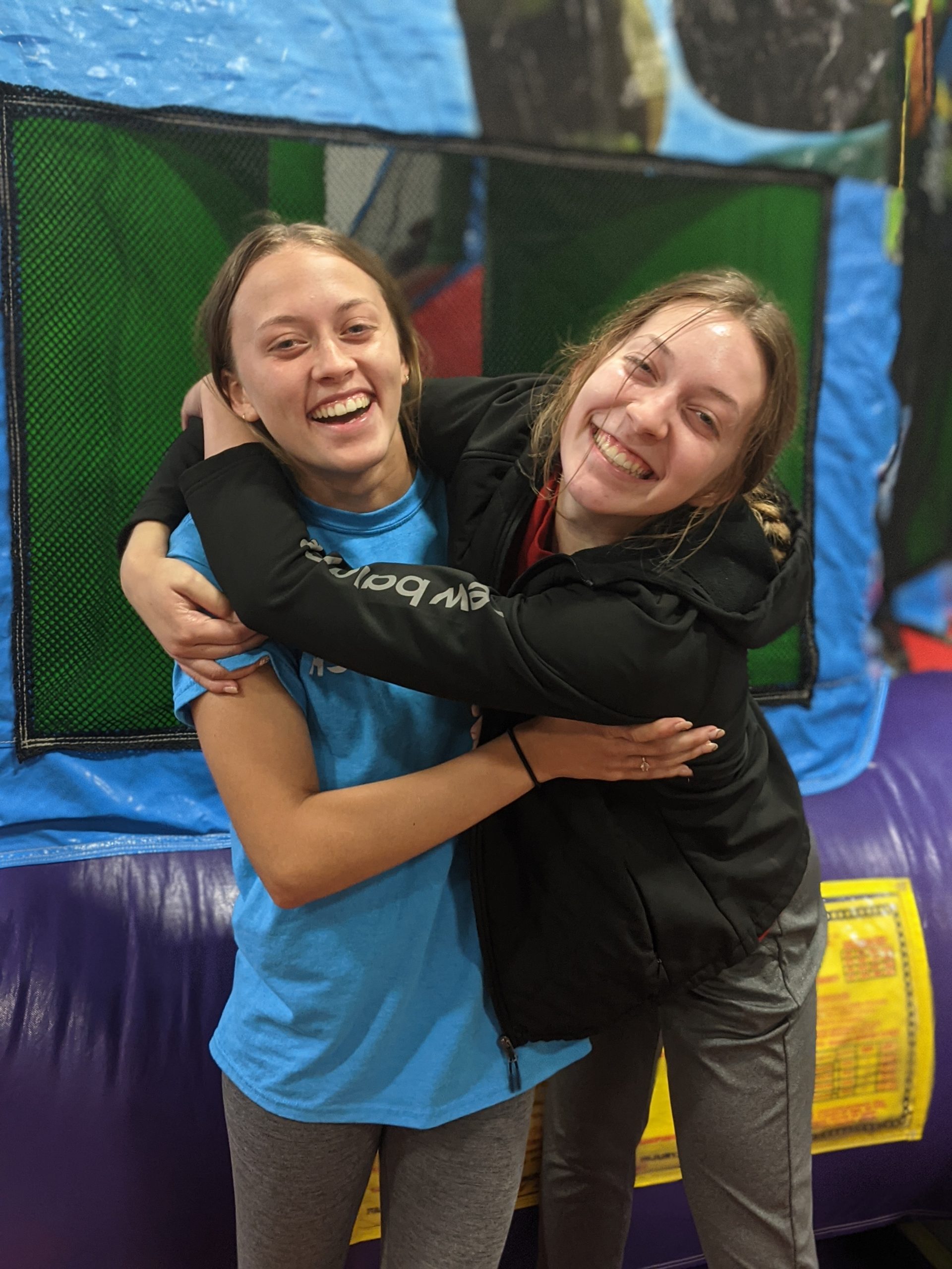 Two friends smiling and hugging in front of a colorful bounce house at an indoor event in Canfield, Ohio, featuring Confetti Event Rental’s safe and fun bounce house rentals popular for parties, schools, and community celebrations throughout Canfield and nearby areas.