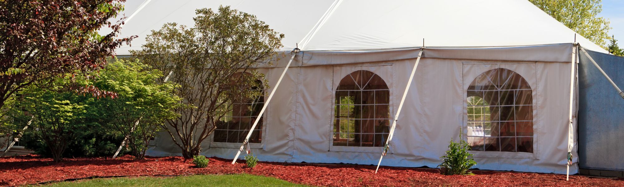White party tent with arched windowed sidewalls beside a red mulch garden bed