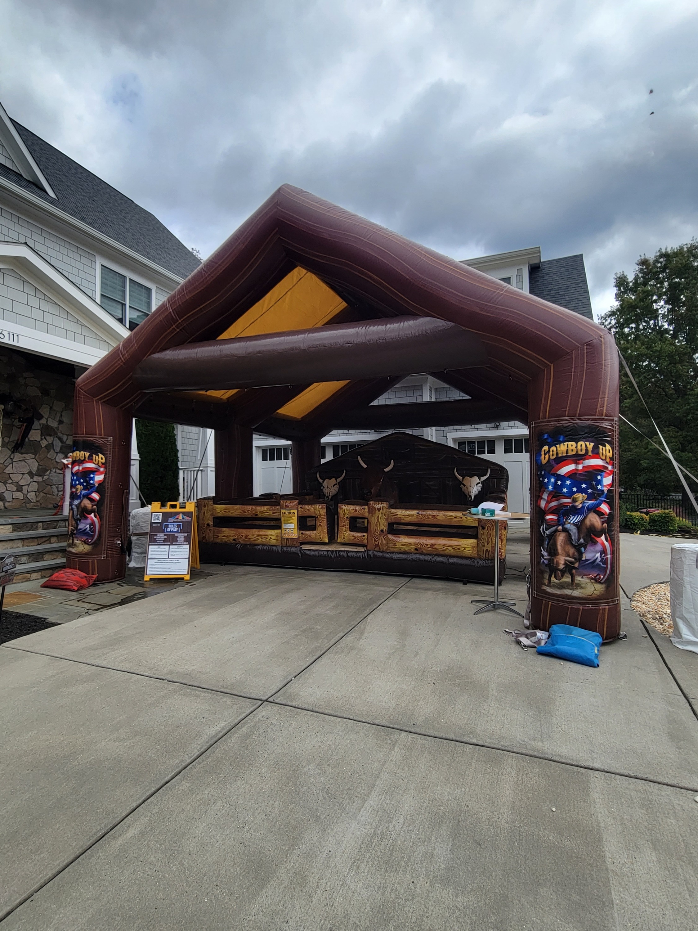 Outdoor setup of a mechanical bull ride by Fiesta Time Amusements in Maryland, enclosed by an inflatable brown canopy with 'COWBOY UP' banners, caution and rules signage, against a backdrop of cloudy skies and residential buildings.