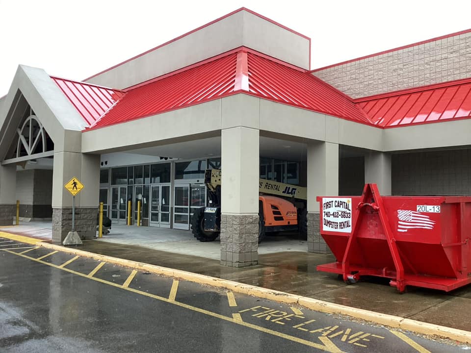 Red dumpster labeled "First Capital Dumpster Rental" outside a commercial building under a red roof. Red dumpster labeled "First Capital Dumpster Rental" outside a commercial building under a red roof.