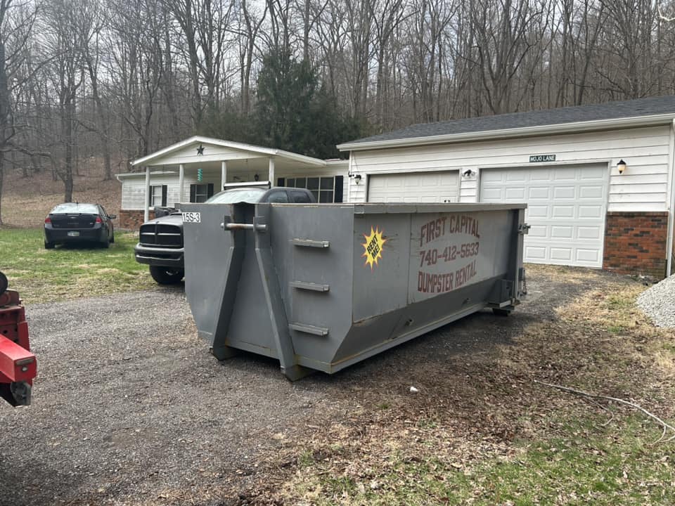 Gray dumpster container labeled "First Capital Dumpster Rental" placed in a residential driveway. Gray dumpster container labeled "First Capital Dumpster Rental" placed in a residential driveway.