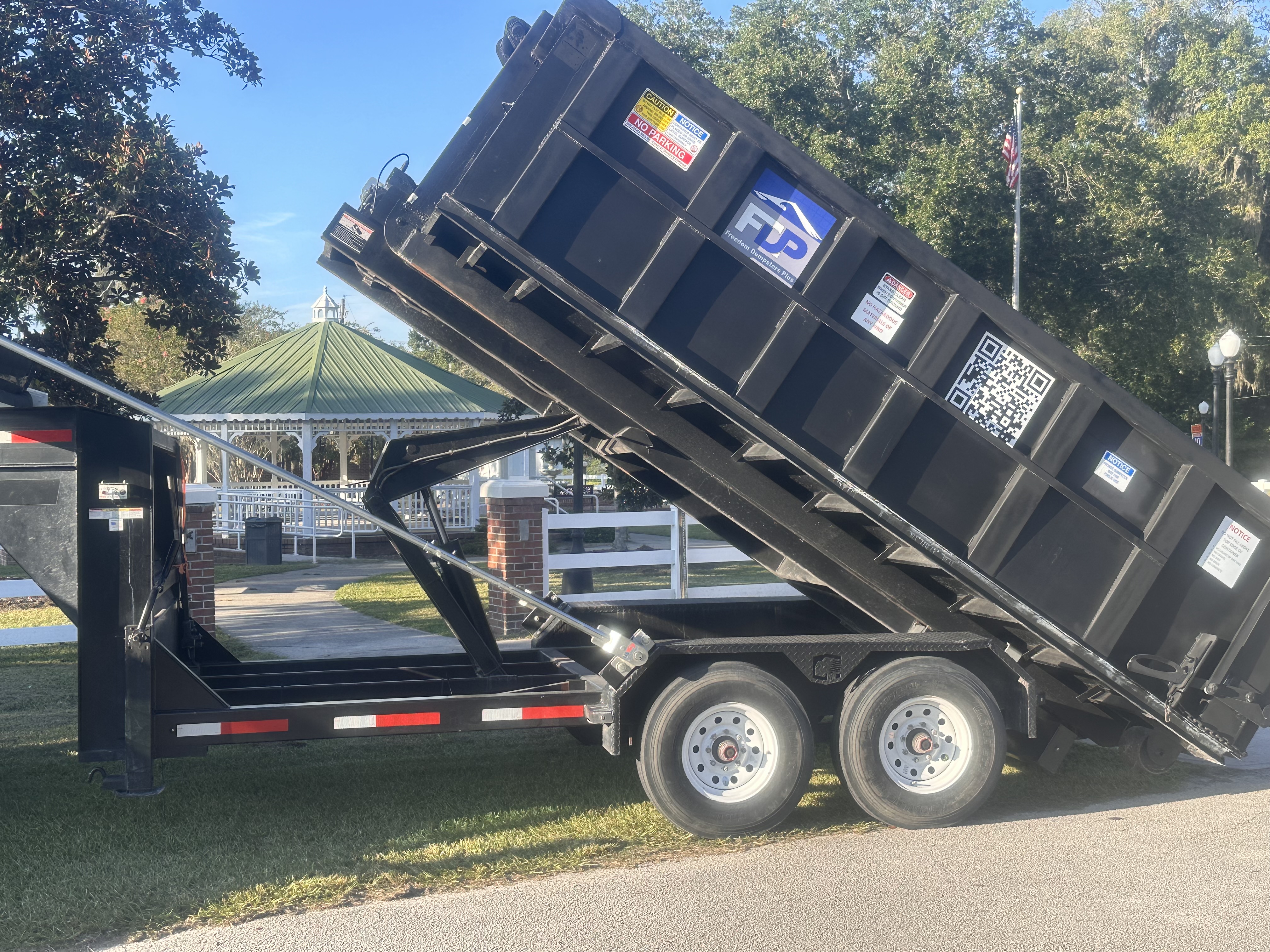 Freedom Dumpsters Plus black roll-off dumpster being unloaded at public park area. Freedom Dumpsters Plus black roll-off dumpster being unloaded at public park area.