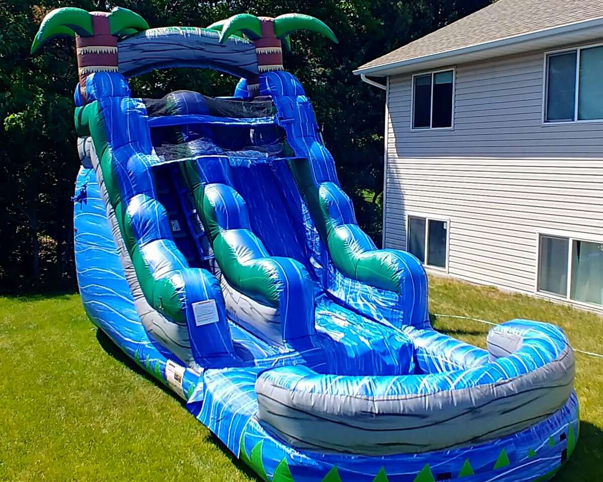 Inflatable blue water slide with palm tree decorations on a grassy lawn beside a house. It