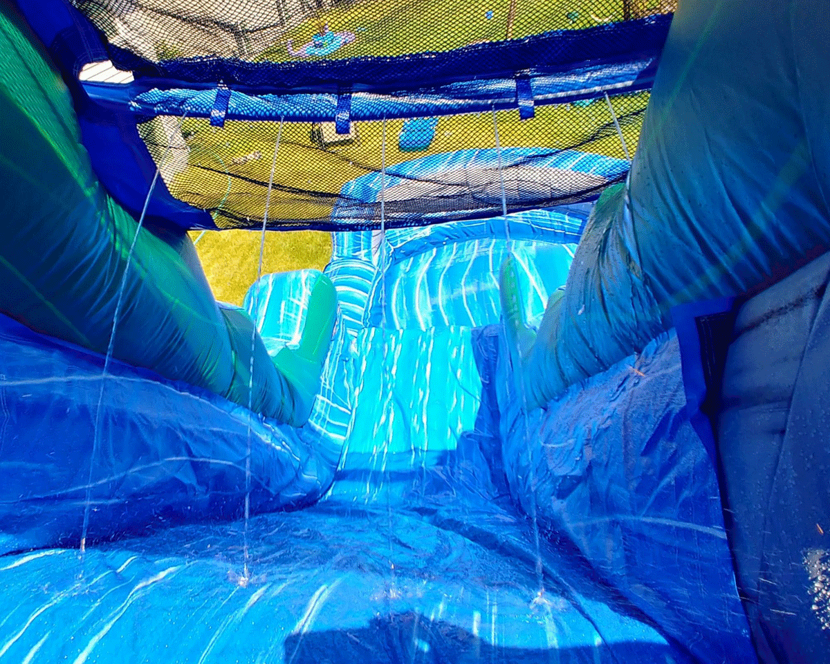 View from the top of a blue inflatable water slide with water spraying down and grass visible at the bottom.
