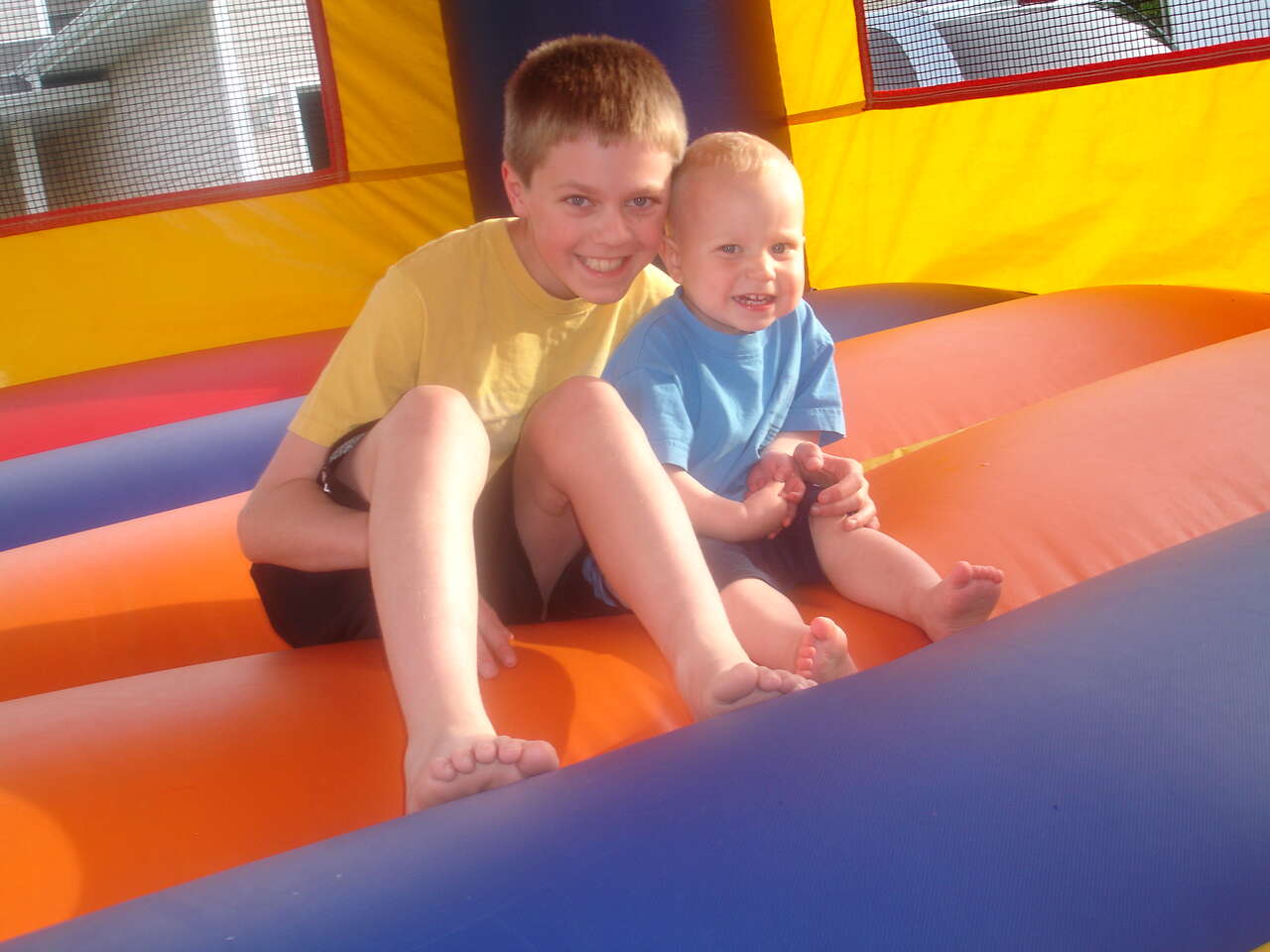 Two young children smiling and sitting inside a colorful inflatable bounce house during an outdoor party or family event.