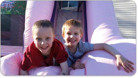 Two smiling children are playing on a pink inflatable slide under a sunny sky. Their expressions convey joy and excitement.