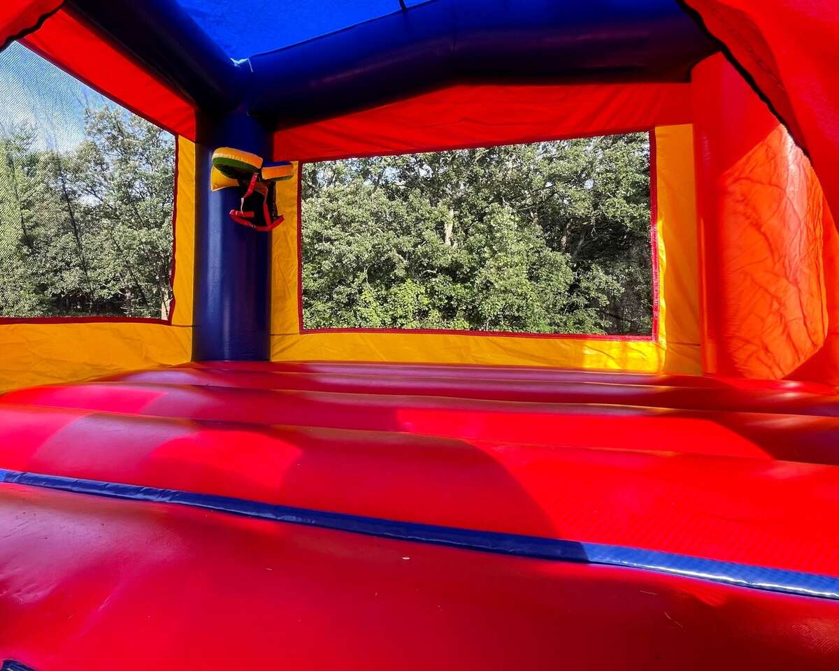 The interior of an empty red, yellow, and blue inflatable bounce house with open windows, set outdoors with trees visible through the windows.