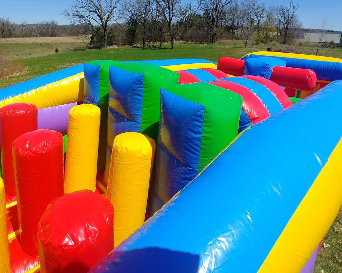 Colorful inflatable obstacle course with blue, red, yellow, and green structures on a grassy field. Trees and a clear sky in the background. Playful and fun atmosphere.