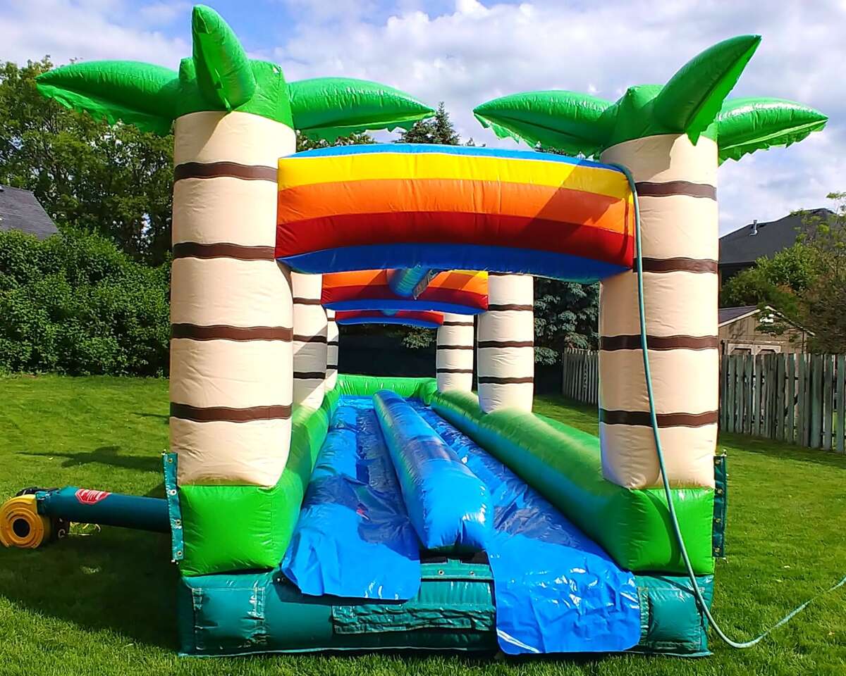 Inflatable water slide with palm tree pillars and a vibrant rainbow arch on a grassy lawn. The scene conveys a fun and playful summer vibe.