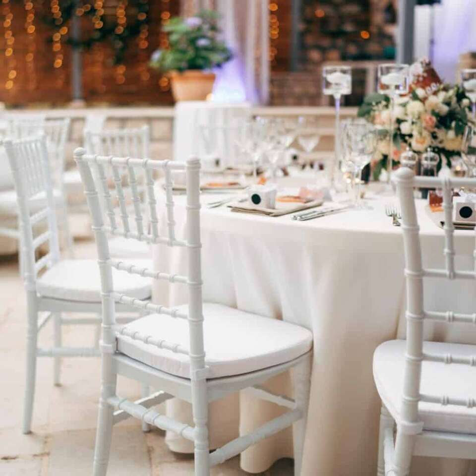 Elegant wedding reception table with white Chiavari chairs, floral centerpieces, candle holders, and warm string lights in a romantic indoor venue setting.