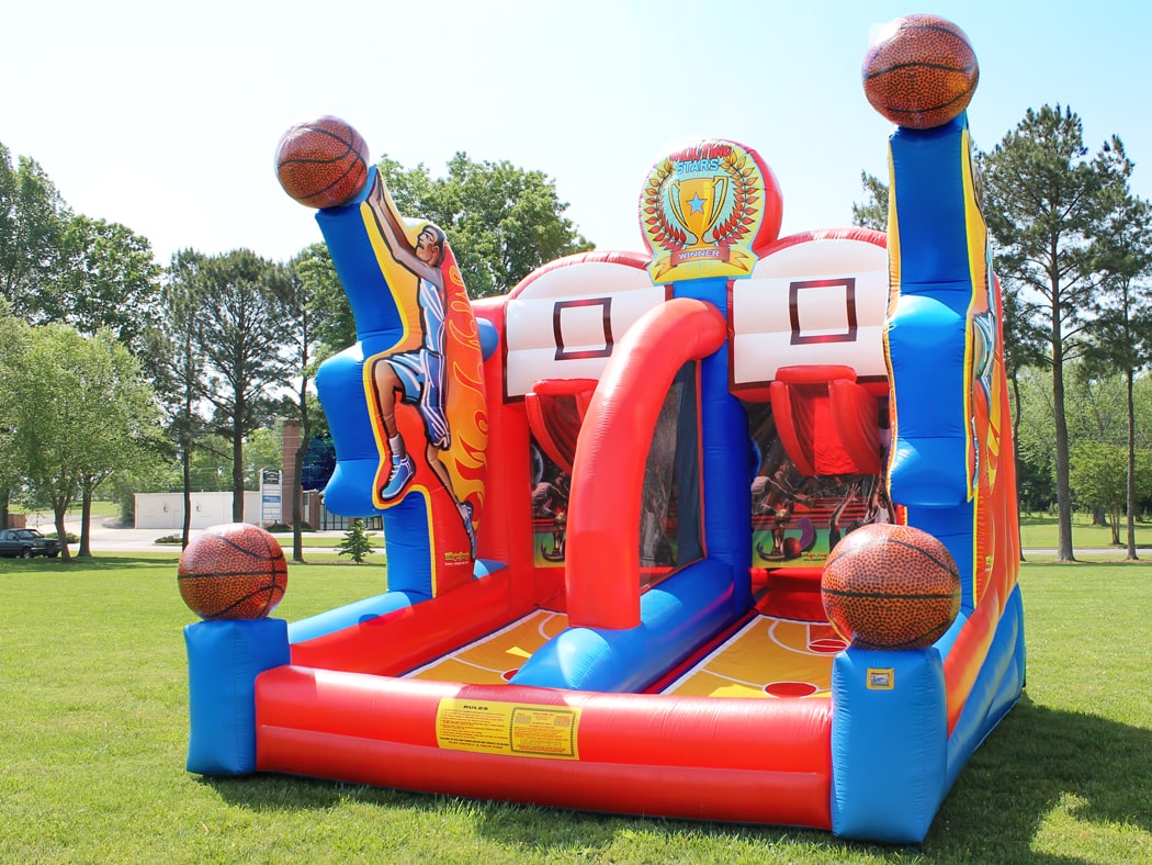 A lively and colorful Shooting Stars Basketball Shootout inflatable game is set up outdoors with two participants ready to play. The inflatable features vibrant red, blue, and yellow colors with large basketball embellishments on each corner, and a backdrop that mimics a basketball court, complete with illustrations of players in action.