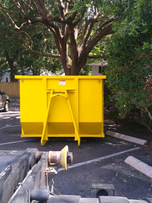 Bright yellow dumpster placed in a parking lot under a large tree.