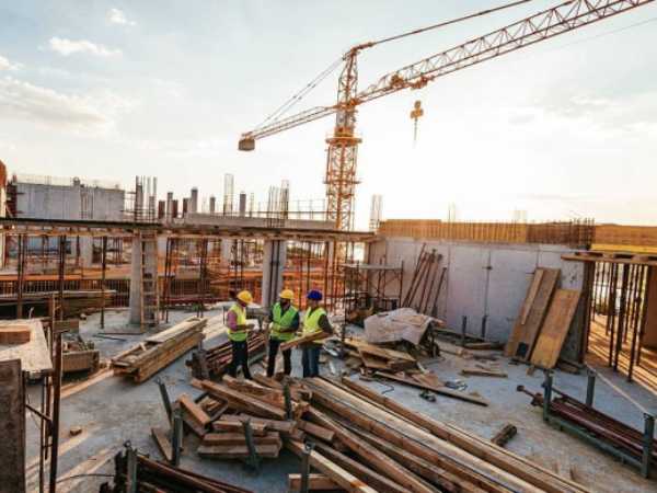 Construction site with workers in safety vests and helmets surrounded by wooden planks and steel bars, with a tower crane in the background at sunset.