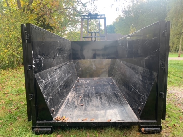 Open-top black metal dumpster placed on grass, showing a clean, empty interior with minor wear marks and scattered leaves at the bottom.