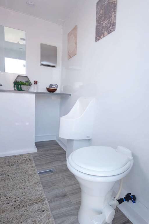 Interior of a luxury restroom trailer rental with a flushing toilet, urinal, running water sink, vanity, and premium finishes for weddings, corporate events, and outdoor events in Buckeye, Arizona